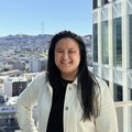 person with long black hair, smiling, standing on a balcony overlooking the skyline of San Francisco