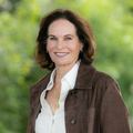 Woman with medium length brown hair wearing a white shirt and brown jacket standing in front of a green backdrop.