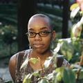 Un joven afroamericano con la cabeza rapada y gafas ovaladas sonríe con picardía mientras mira a la cámara, con un rosal en primer plano.