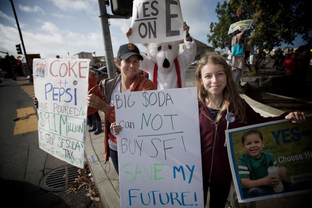 People hold signs supporting San Francisco’s soda tax, including messages like “Big Soda can NOT buy SF!” and “Yes on E.”