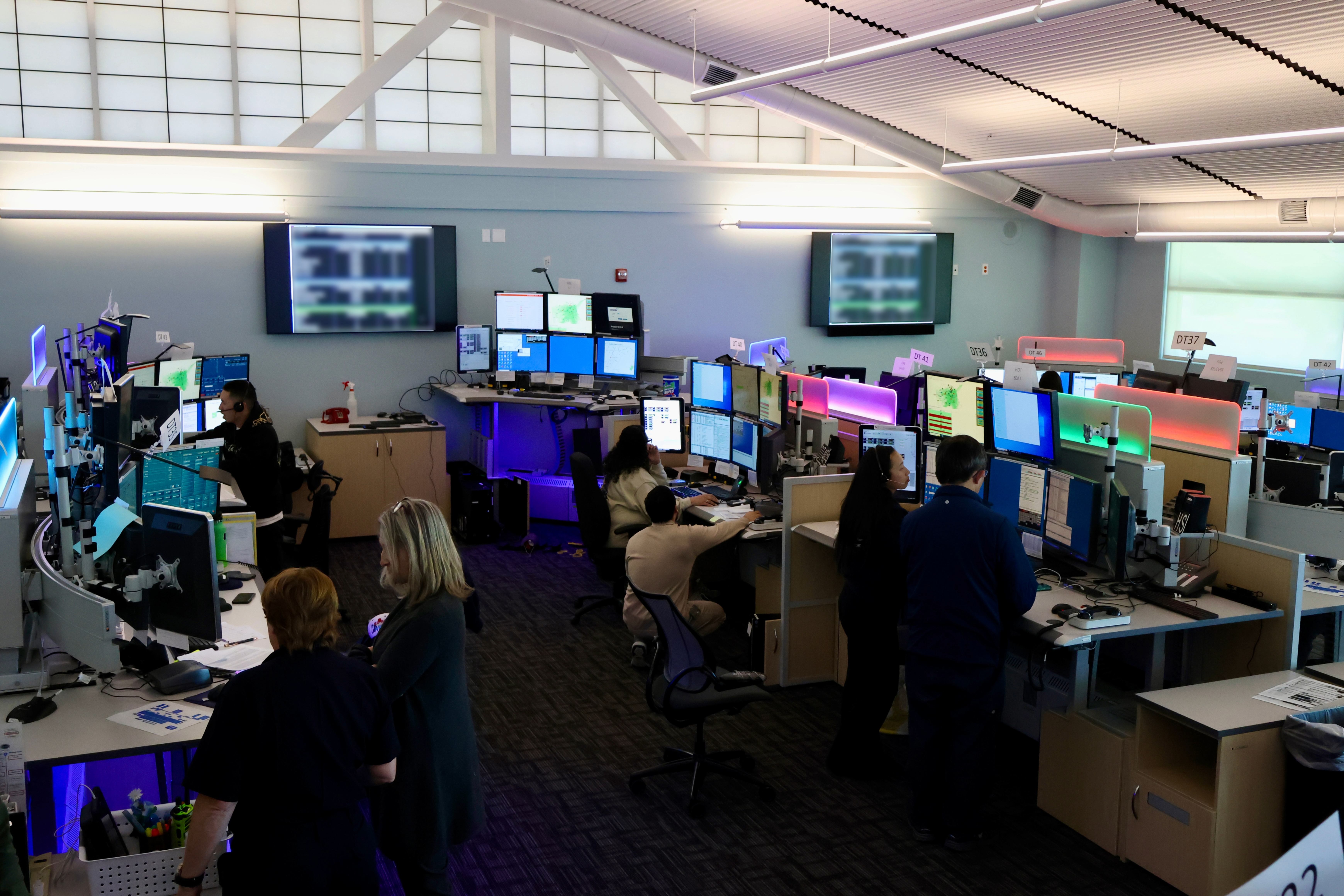 Emergency communications center with dispatchers working at multiple computer stations, large monitors on the wall, and staff collaborating throughout the room.