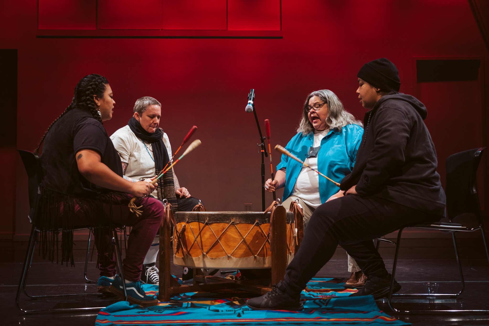 Four performers sit in a circle on stage, drumming and singing together around a large drum, illuminated by warm red lighting