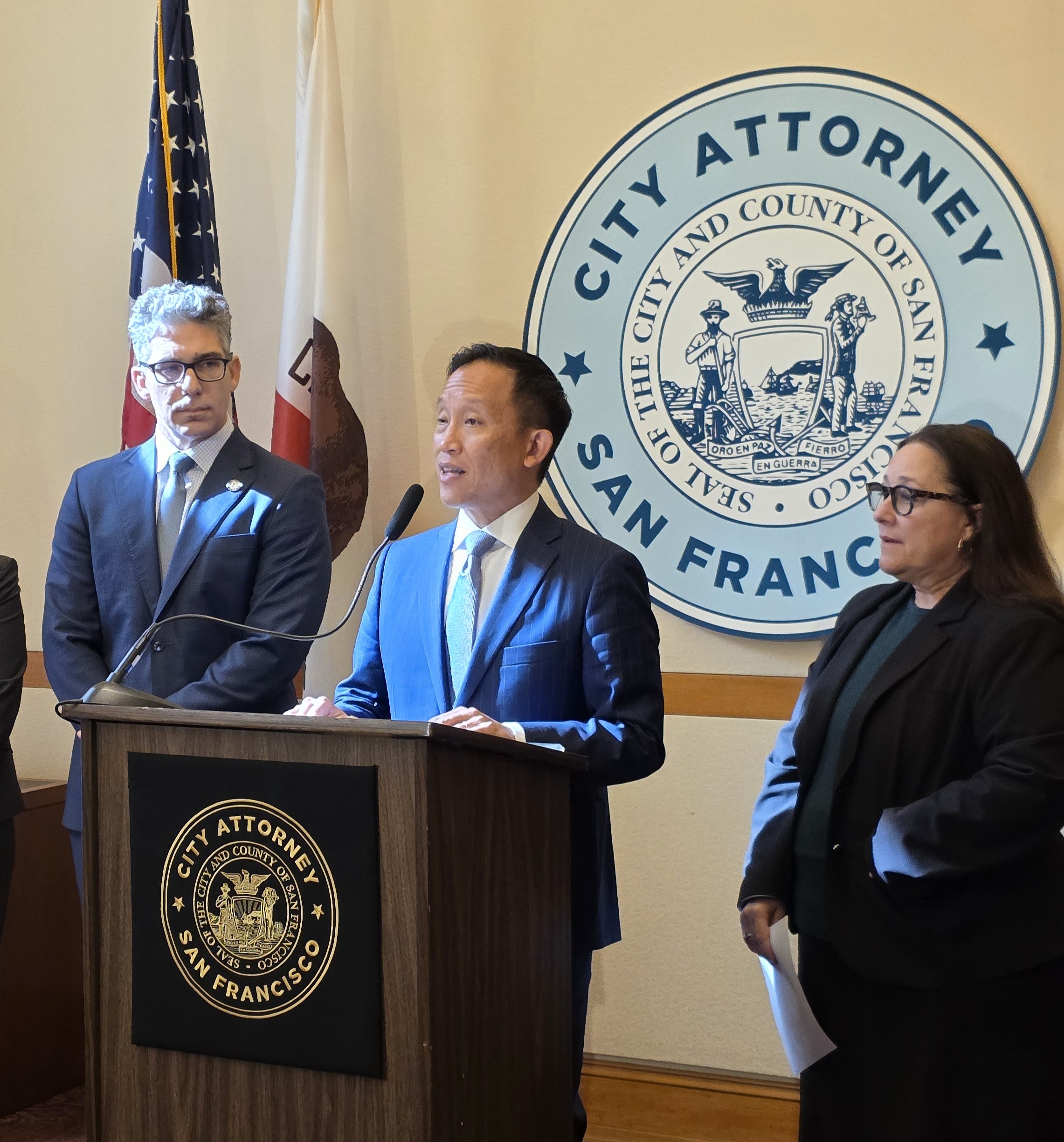 San Francisco City Attorney David Chiu speaks at a podium alongside Santa Clara County Counsel Tony LoPresti and San Francisco Chief Deputy City Attorney Yvonne R. Meré in February 2025
