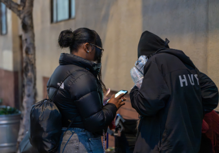 2026 PIT Count_Two people standing on a city sidewalk, one showing something on a smartphone while speaking with another person