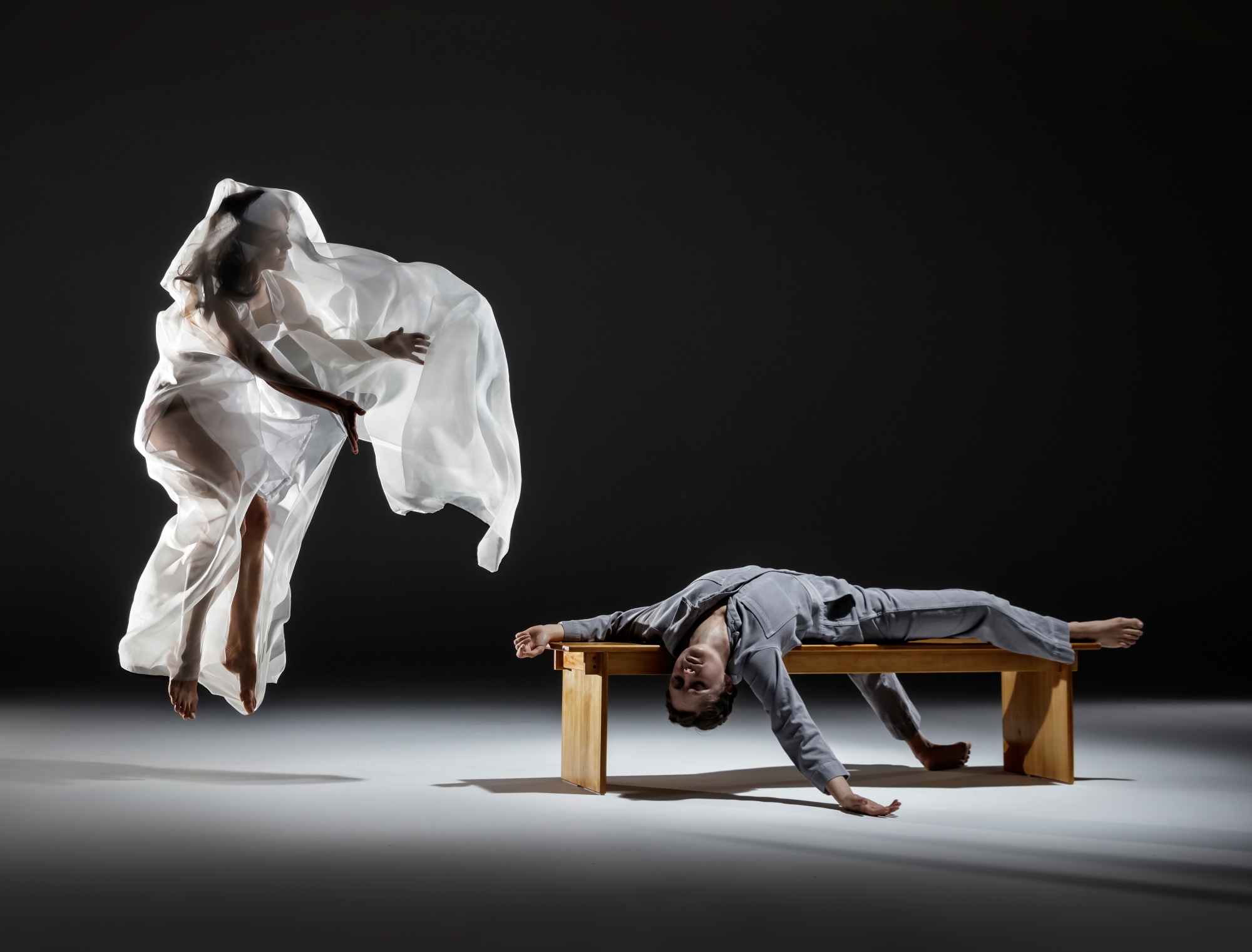 2 dancers, one on the left is veiled in white jumping, the other is in grey splayed out on a brown wooden bench