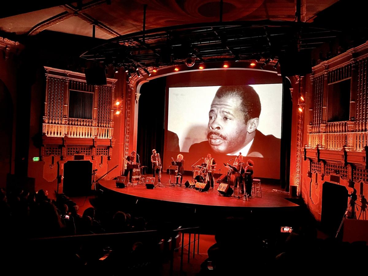 stage with 6 musicians lit by a red light with a backdrop of an african male