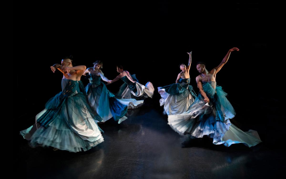 A group of dancers in flowing teal dresses perform gracefully on a dark stage, captured mid-motion with expressive, sweeping movements.