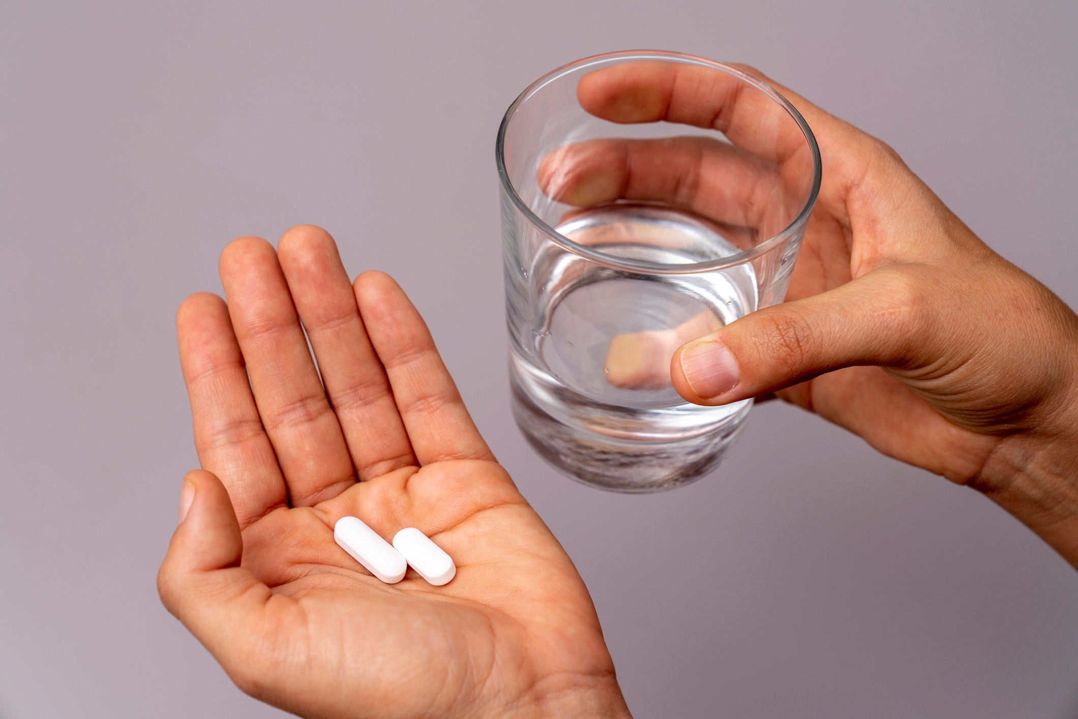 a hand holding 2 white pills and another hand holding a glass of water