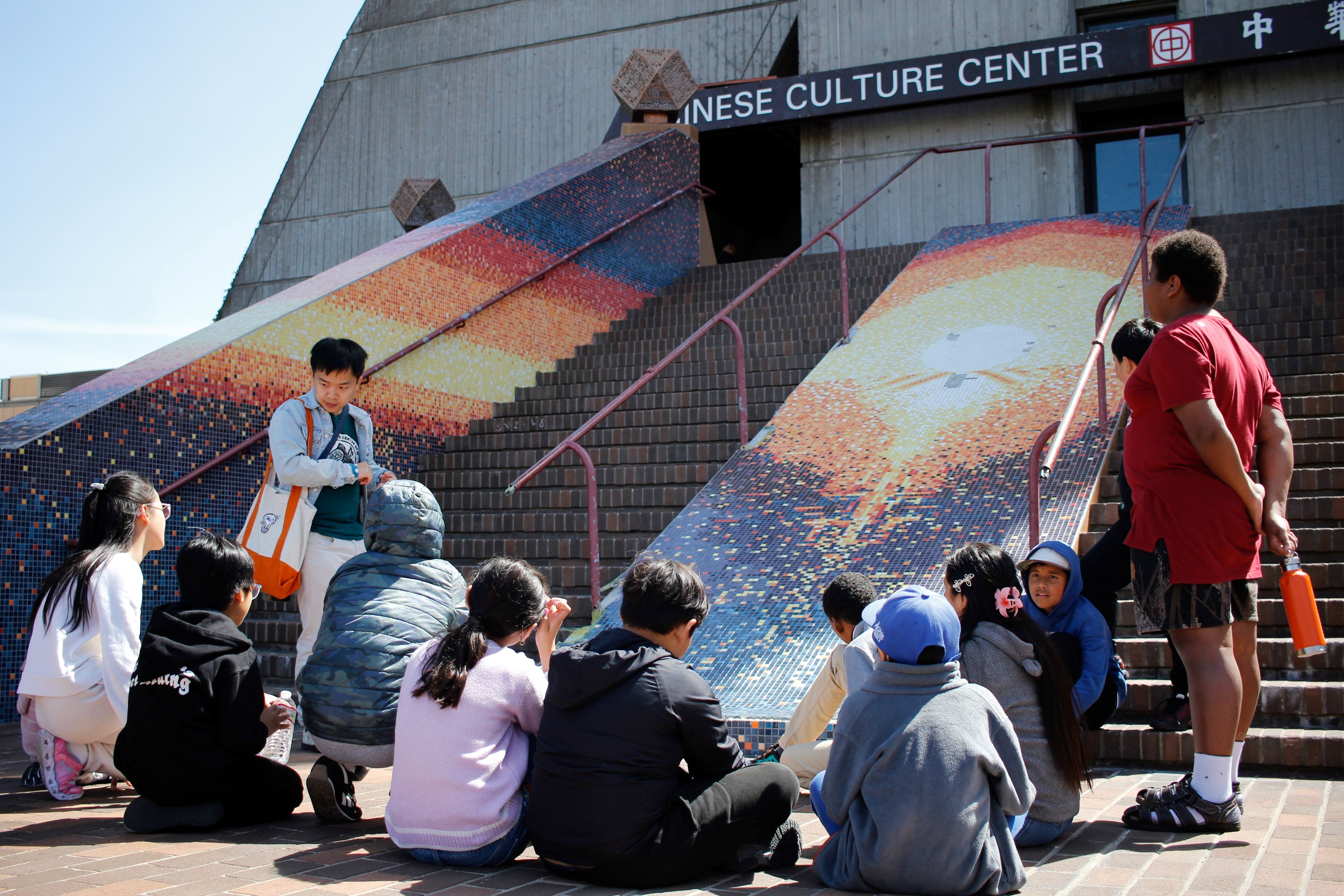 A group of people sits and stands on brick steps outside the Chinese Culture Center, listening near two large, colorful mosaic ramps leading up to the entrance.