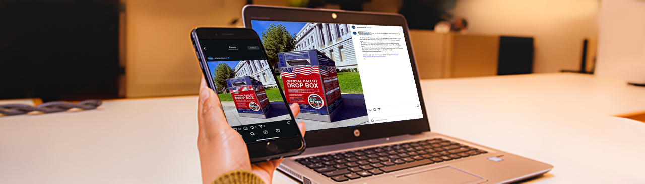 Hand holding cell phone next to laptop looking at the SF Elections social media page.