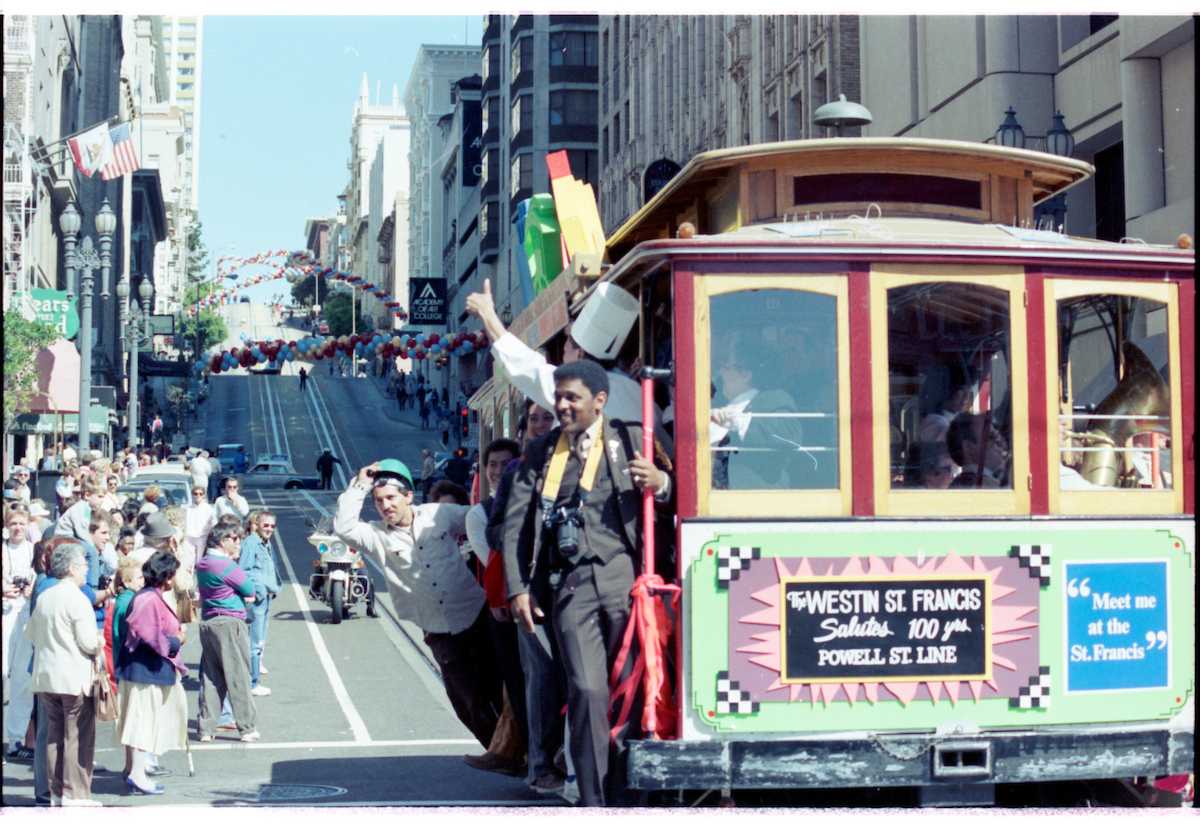 An image from SFMTA's archive showing people standing on a cable car, driving past crowds of people.