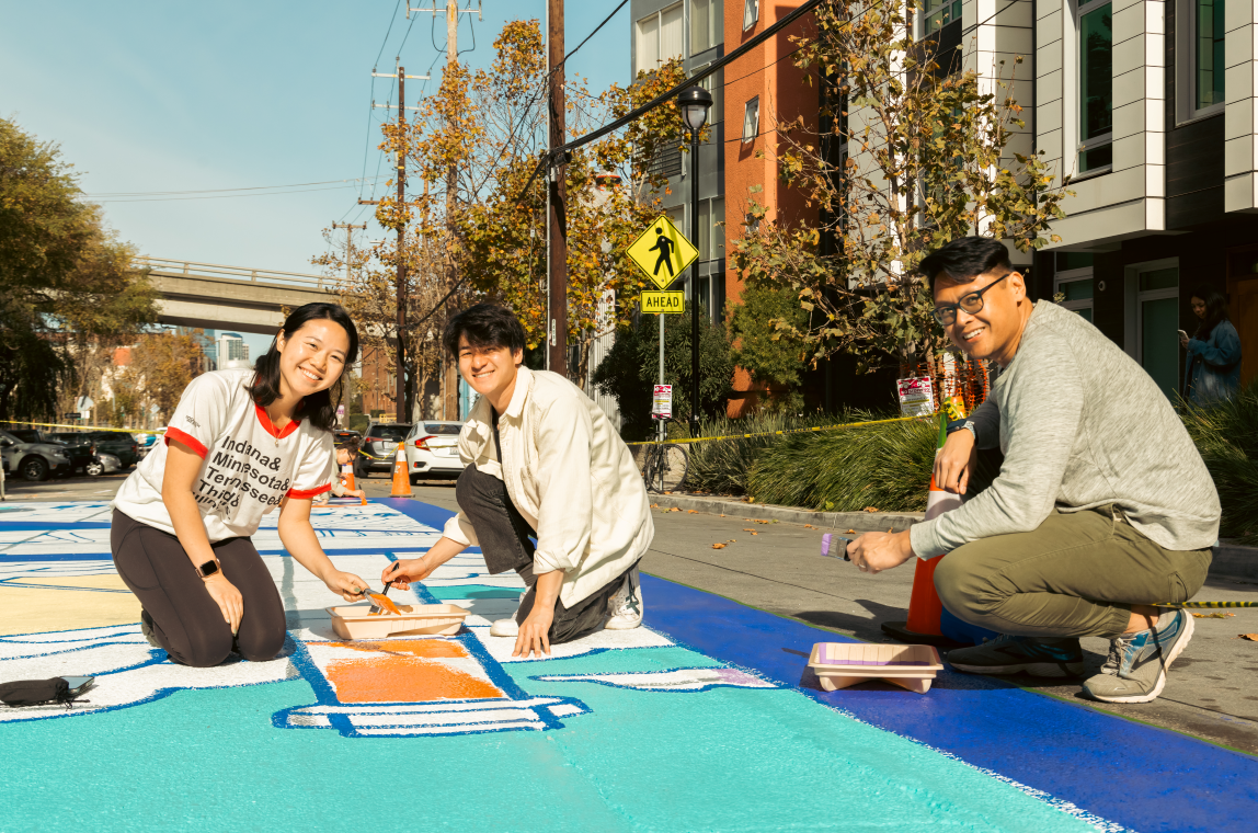 Three people smile while painting a colorful mural on a sunny street. They are surrounded by buildings and autumn trees, creating a cheerful atmosphere.