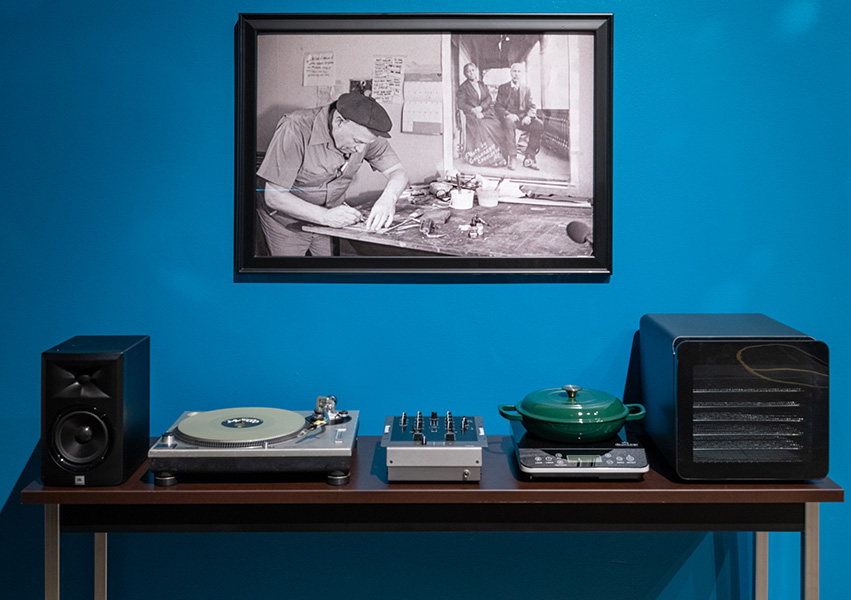 Isang instalasyong sining ng isang mesa na may record player, isang palayok na bakal, at mga speaker. Isang itim at puting litrato ng isang lalaking nakasuot ng jumpsuit na nagtatrabaho ang nakasabit sa dingding.