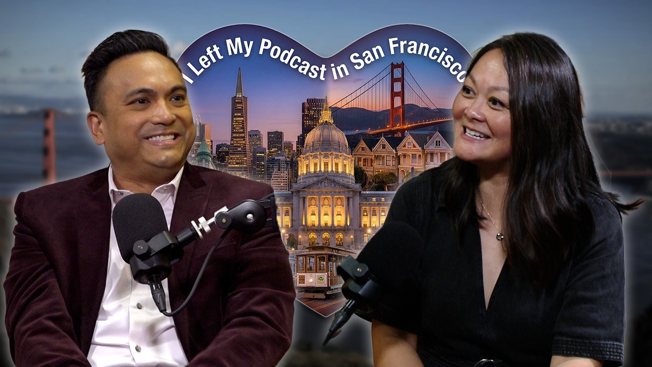 A man in a suit sits in front of a microphone. A dark haired woman in a dress smiles.