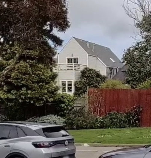 The back of a two-story house visible from across the street through the backyard.