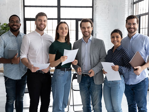 Six people standing in a bright room, smiling and holding papers, in front of large windows.
