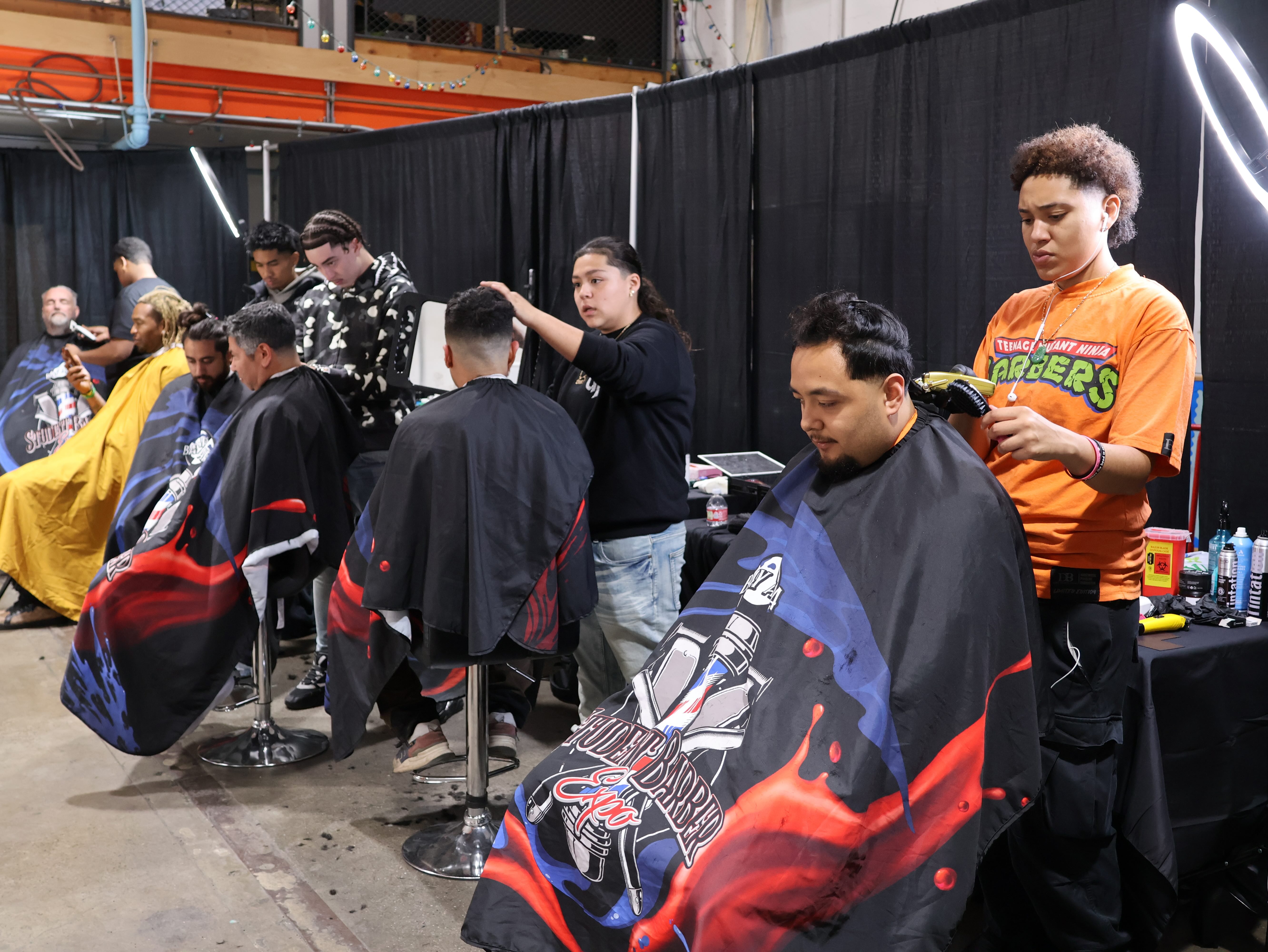 Barber pathway graduates give haircuts to clients seated in colorful barber capes at several workstations.