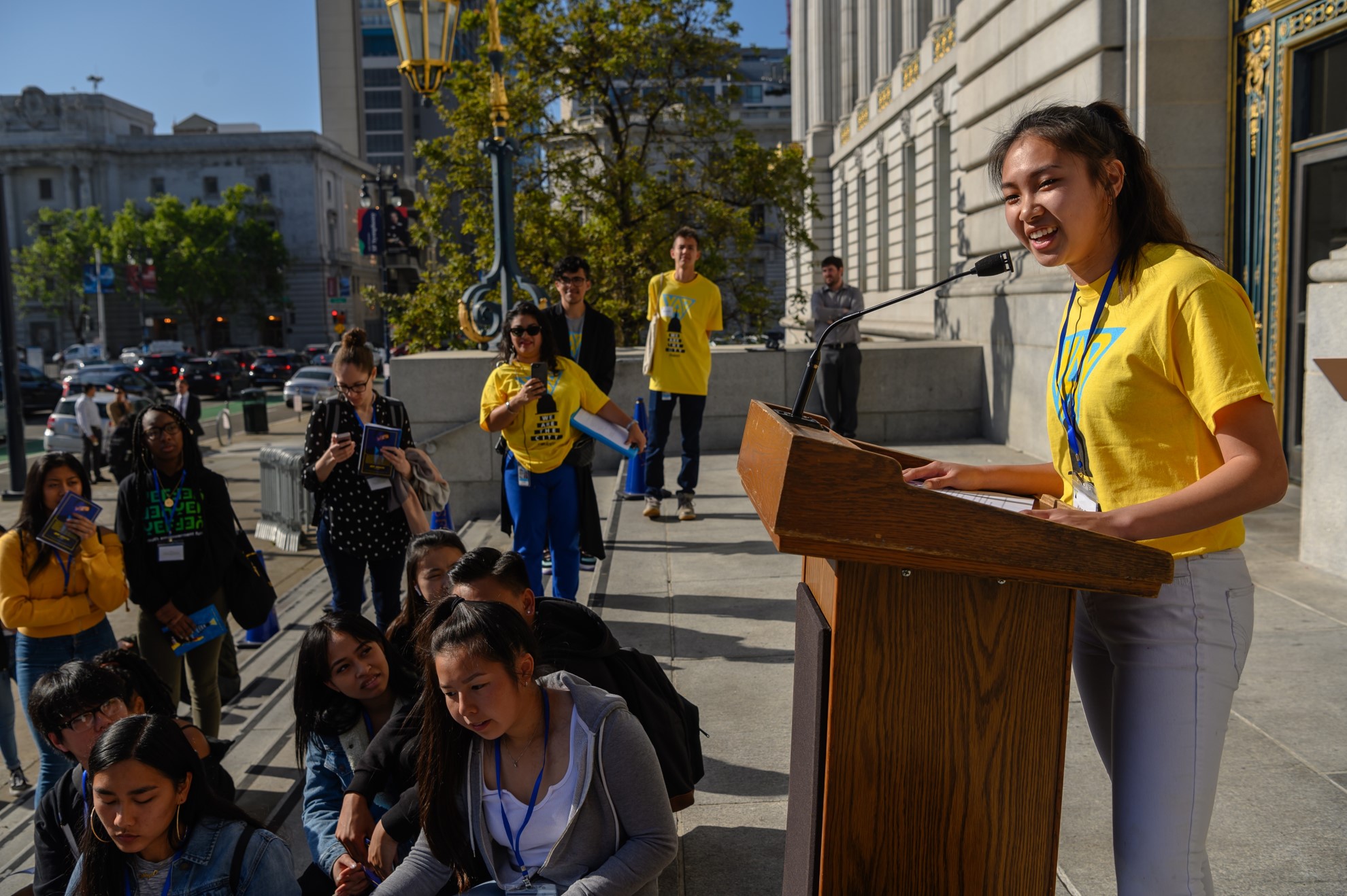 High school girl at a podium speaking to a crowd of youth in front of San Francisco City Hall.