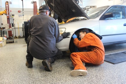 Student working under a car while an instructor watches.