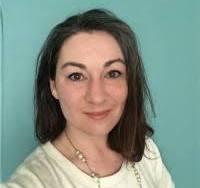 A photo of a white woman with dark brown shoulder length hair is in a white shirt in front of a teal wall