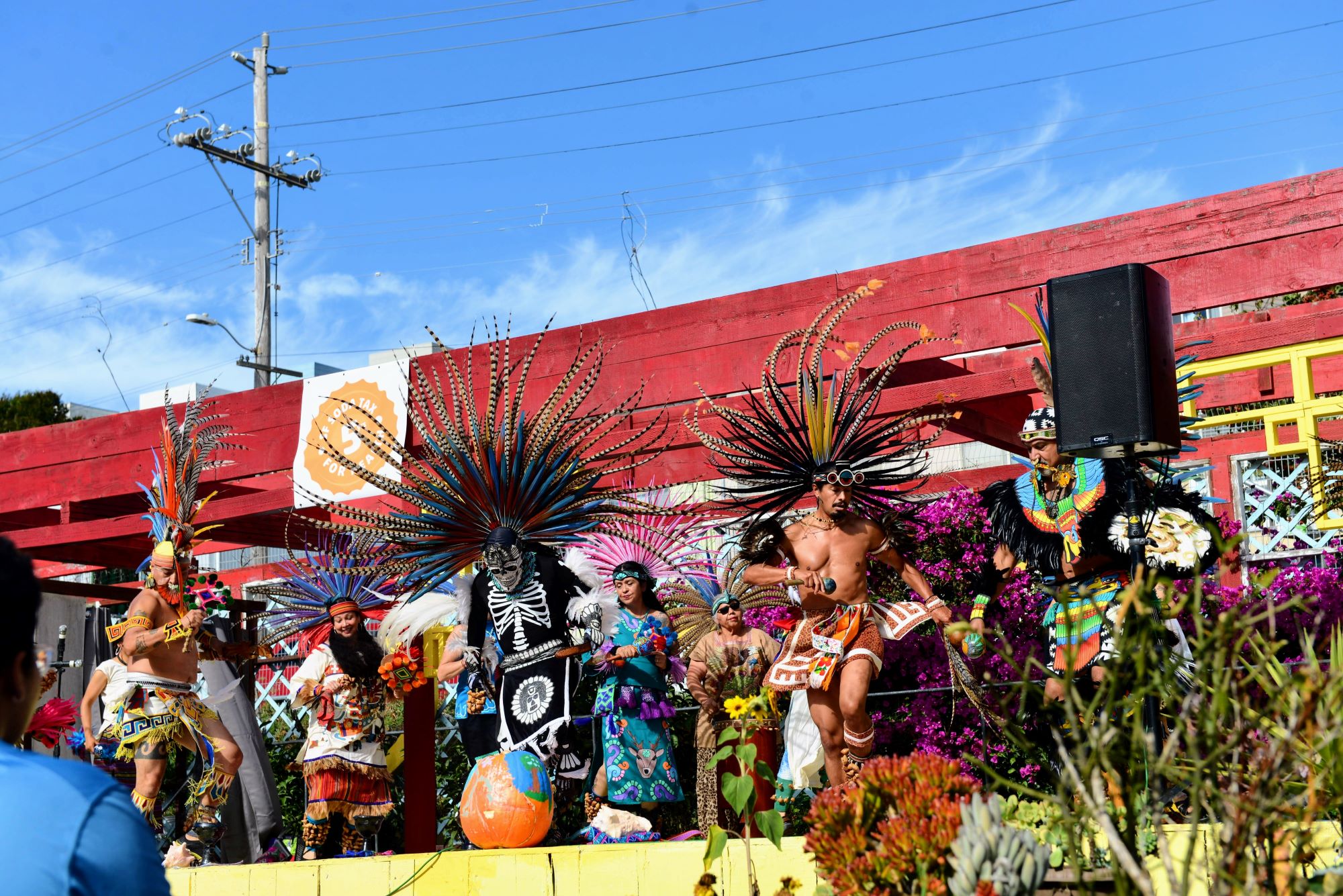 Dancers in colorful traditional Danza Azteca Mexica Indigenous regalia with feathered headdresses perform outdoors before a red stage backdrop.