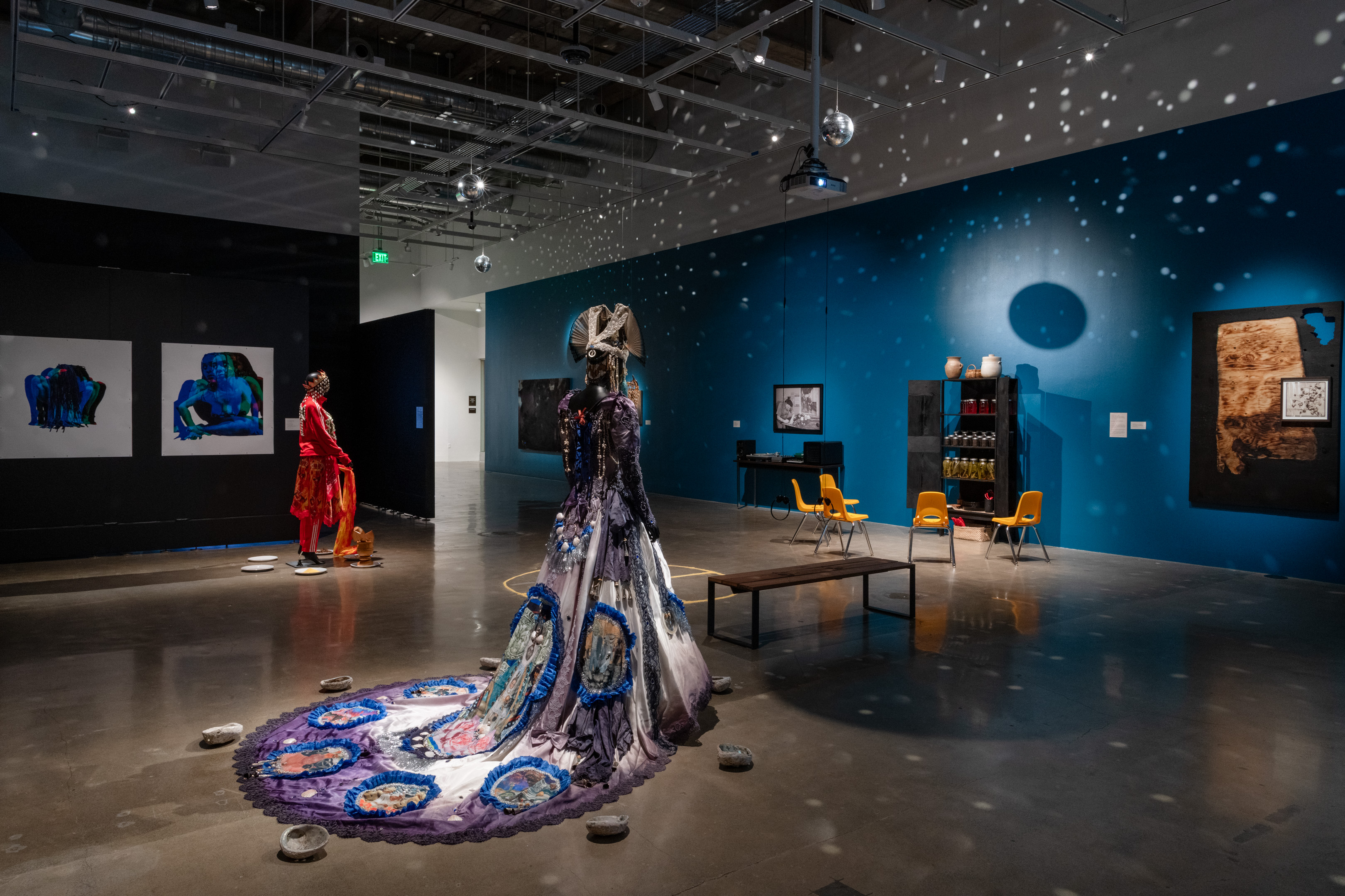 A contemporary art installation in a gallery: textile works on mannequins, sculptural installations, and projected lights.