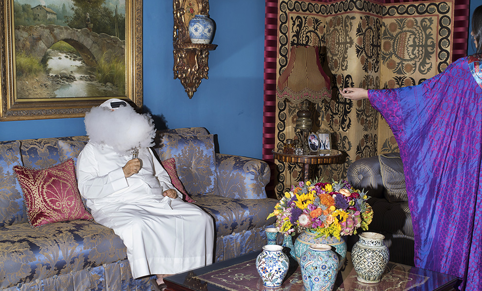 Man seated on an ornate couch in a richly decorated living room, his face obscured by a cloud of vape smoke, with patterned textiles and decorative objects surrounding him.