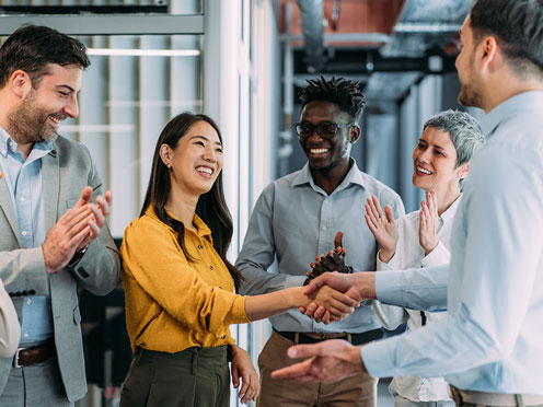 A group of five people, smiling and applauding, with two individuals shaking hands in an office setting.