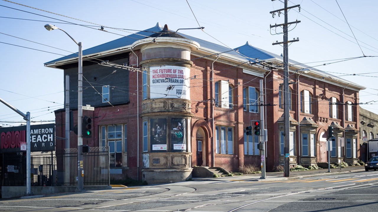 red brick building with posters adorning the corner of the building as well as some windows. Powerlines run across the top half of the frame. There is an empty street.