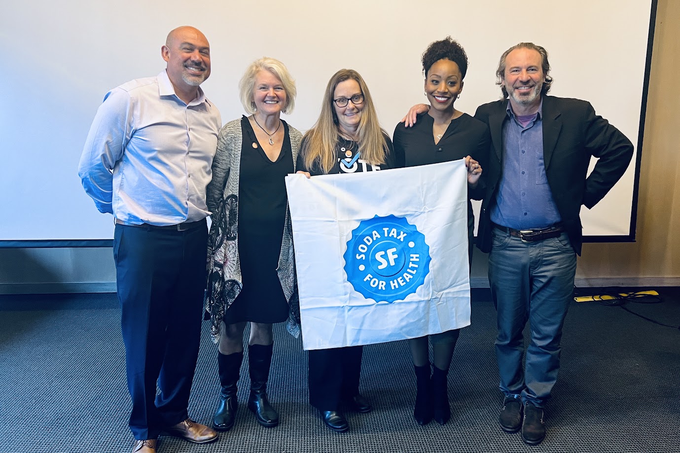 A group of five people standing together in a lecture hall, smiling and holding a white banner with a blue logo that reads “SF Soda Tax for Health.”