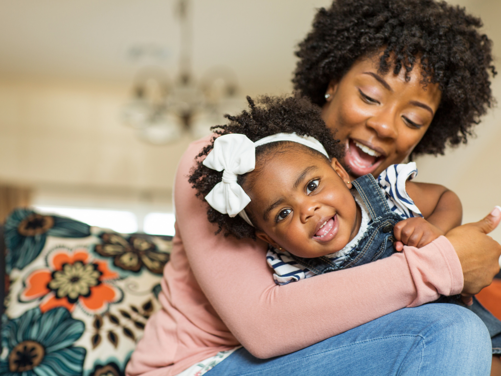 A woman is hugging a young child. Both are smiling and looking at the camera. The child has a white bow in her hair.