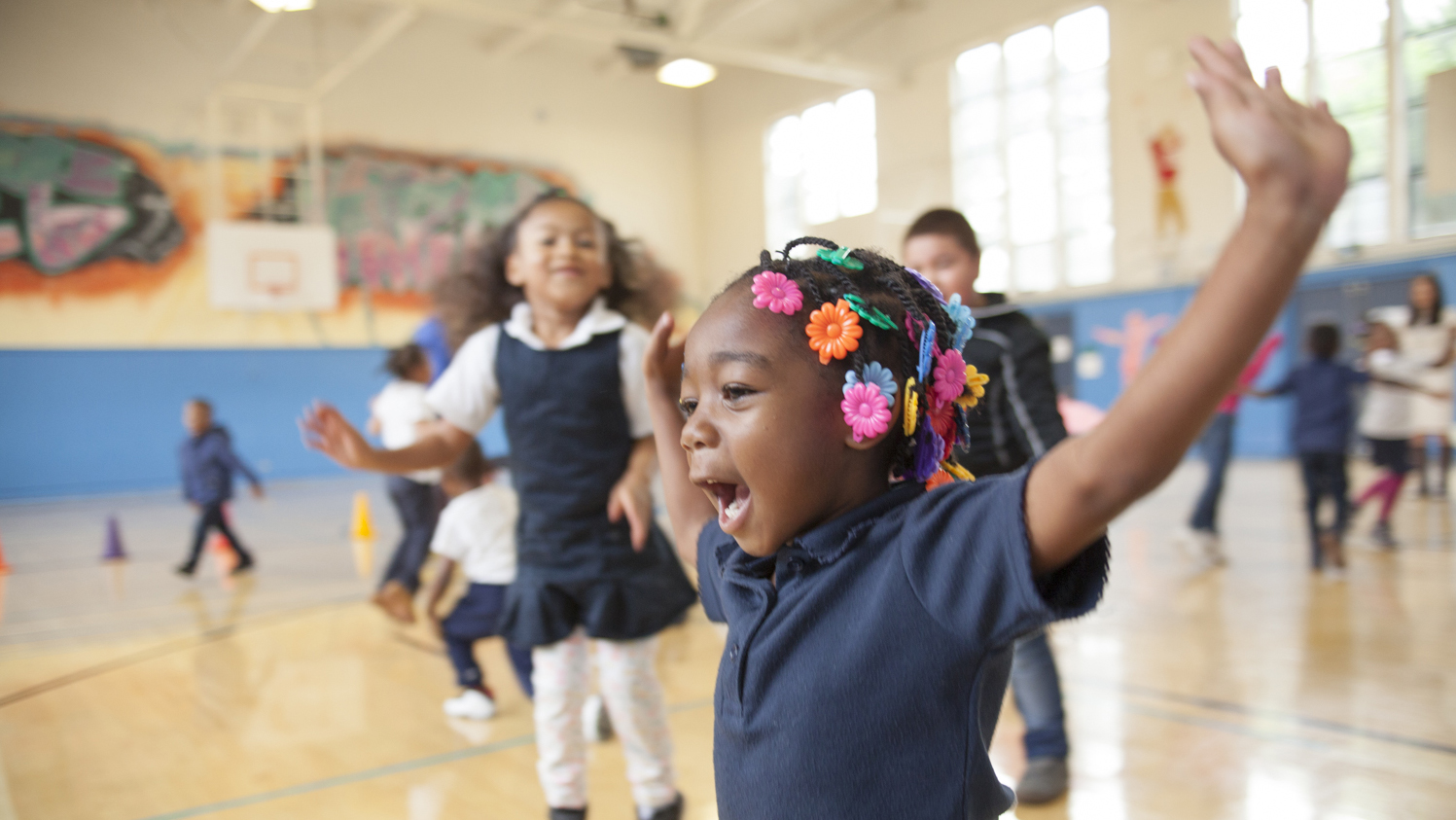 Smiling young girl with arms up inside a gym. Other children are visible doing physical activity in the background.