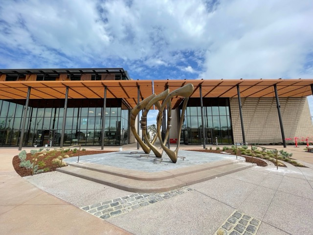 Modern building with glass façade and wood canopy, featuring a large abstract metal sculpture in the plaza.