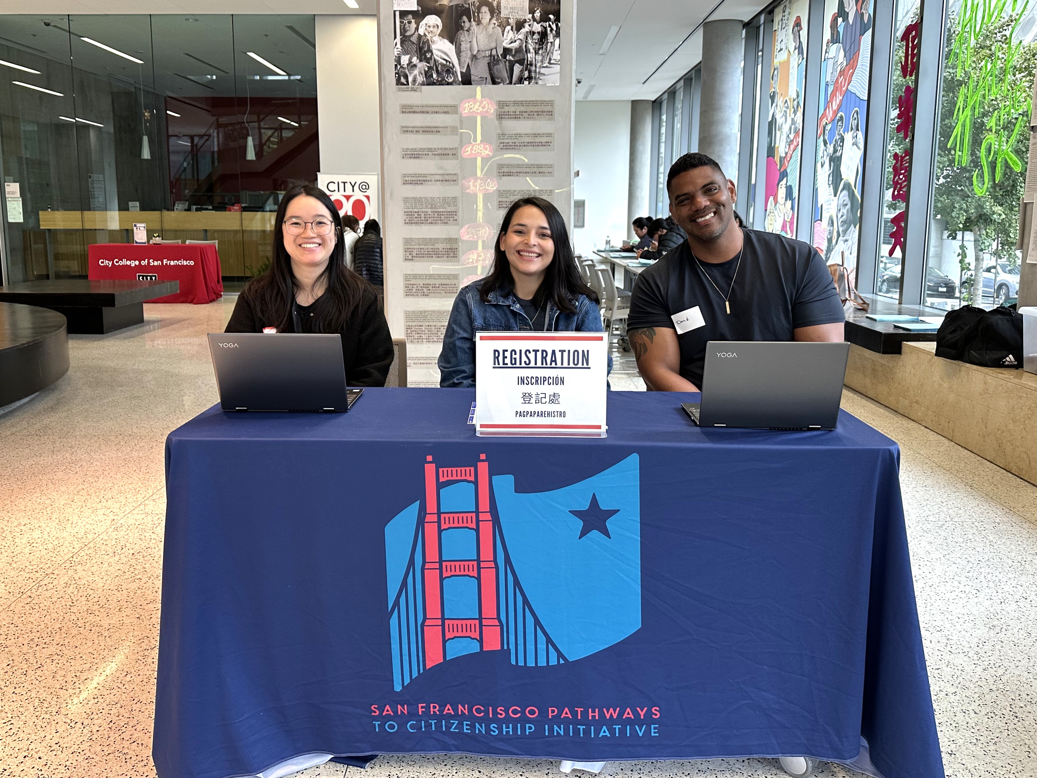 Three SF Pathways to Citizenship volunteers smile at the registration table at a citizenship workshop, welcoming participants.