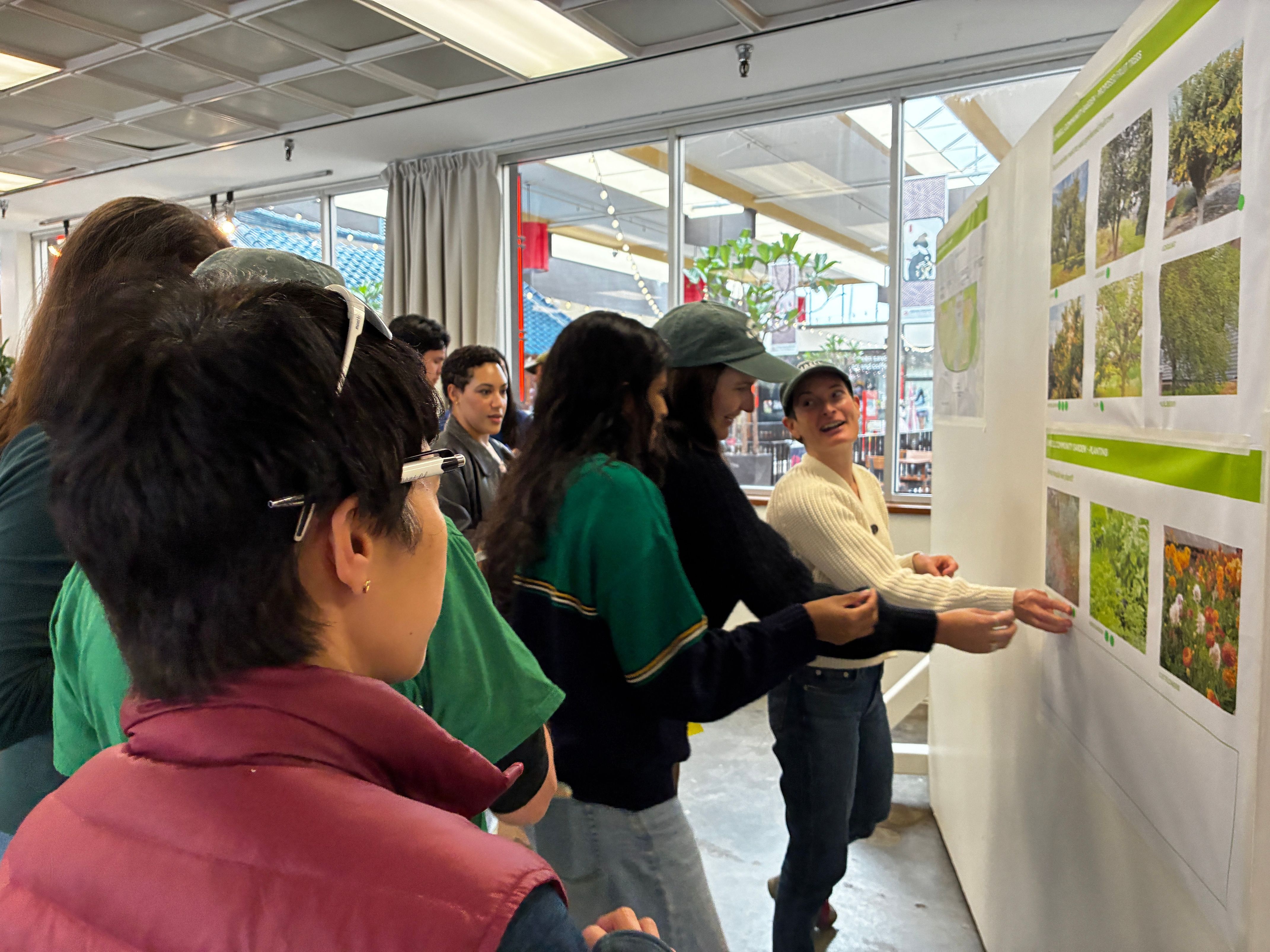 People gathered indoors look at and gesture toward a display board with printed photos of plants and outdoor landscapes.