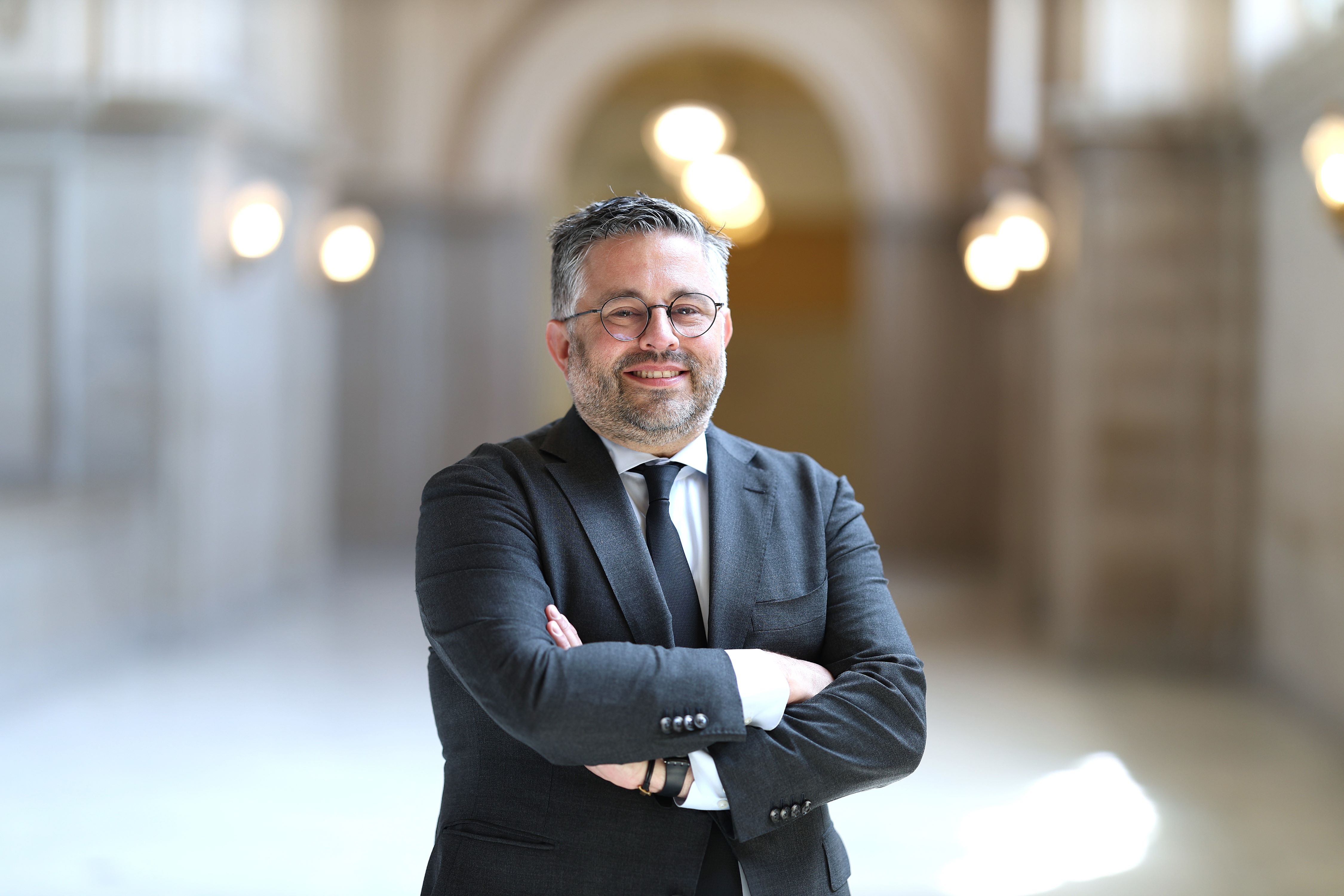 Man wearing glasses and a dark suit and tie, standing in a hallway in San Francisco City Hall