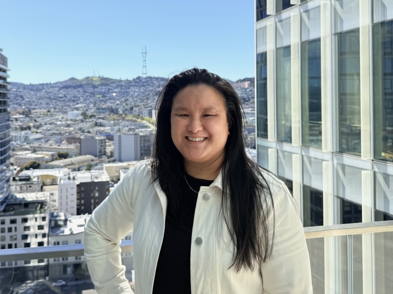 person with long black hair, smiling, standing on a balcony overlooking the skyline of San Francisco