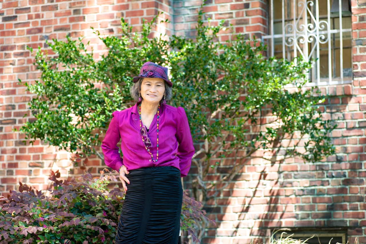 Asian American trans woman wearing a lavender colored blouse, matching hat and black skirt posing in front of a small tree