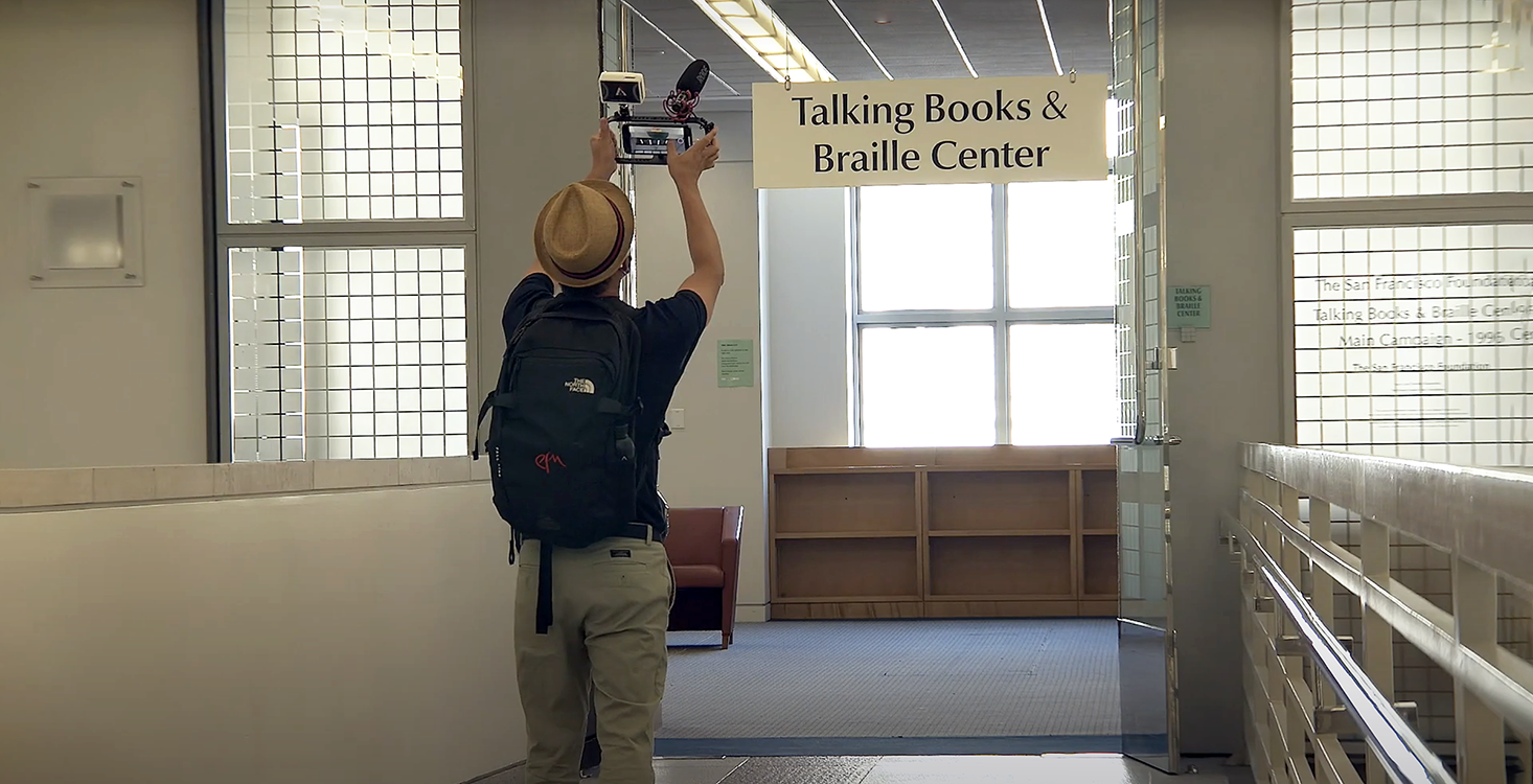 A person wearing a hat and a backpack., holding up a video camera filming an open door with a sign above it stating "Talking Books and Braille Center."