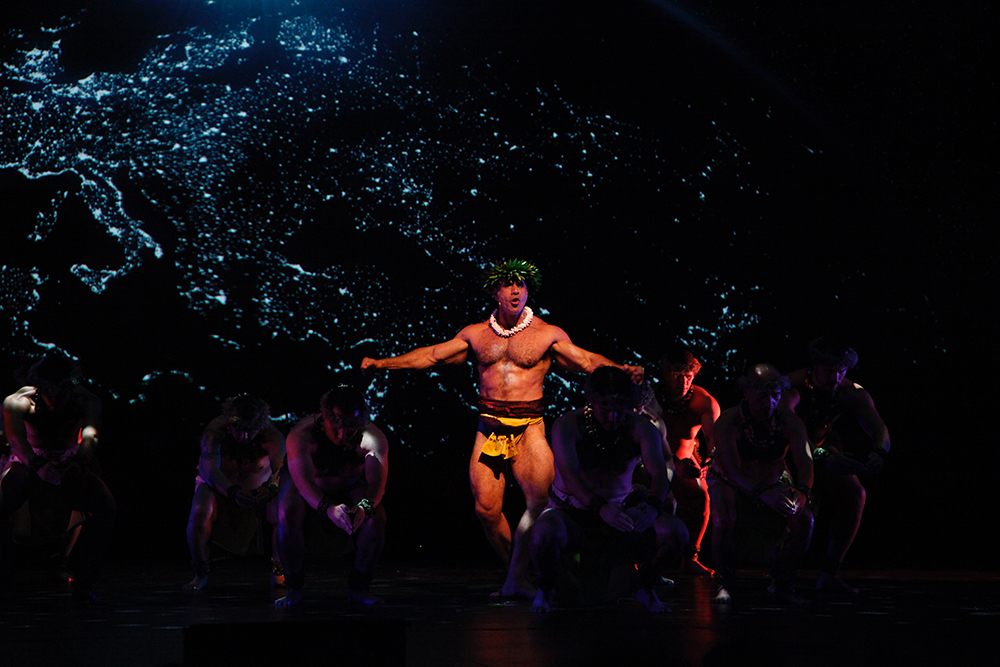 A powerful stage performance features dancers in traditional attire against a glowing, map-like backdrop, evoking cultural identity and connection.