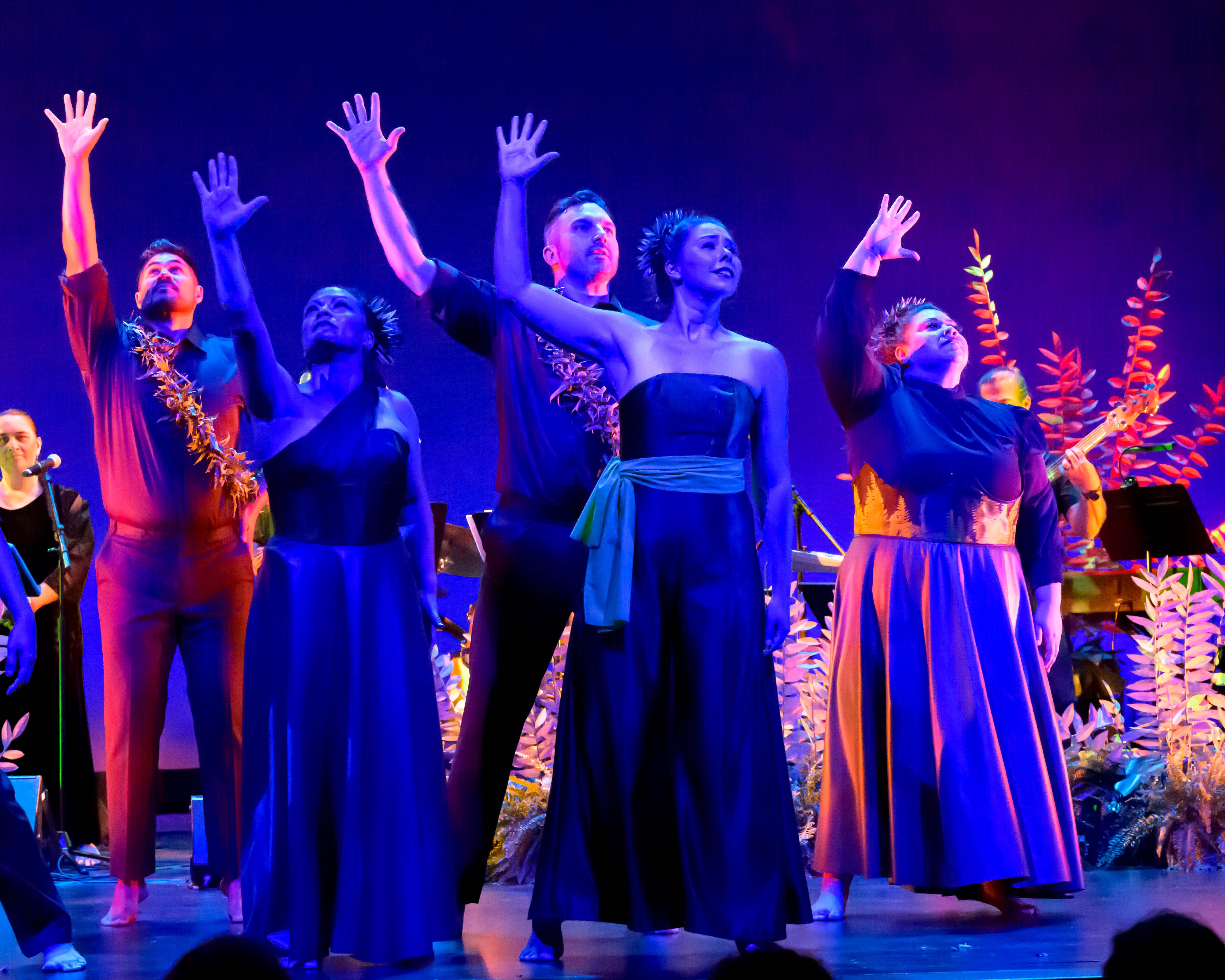 Five performers stand on a stage under blue and purple lighting, dressed in flowing, earth-toned costumes with leaf adornments. They raise one arm upward in unison, gazing toward the audience, while musicians and decorative foliage are visible in the background, creating a dramatic, nature-inspired performance scene.