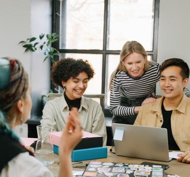 Four people gathered around a table with a laptop and office supplies, engaged in a collaborative discussion.