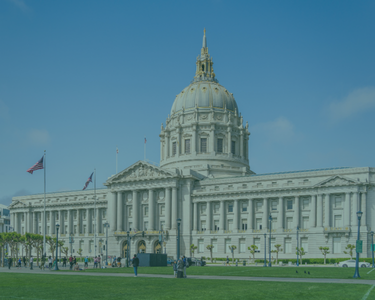 Exterior view of San Francisco City Hall under a clear sky, showing the large dome, columns, flags, and people walking across the lawn in front of the building.