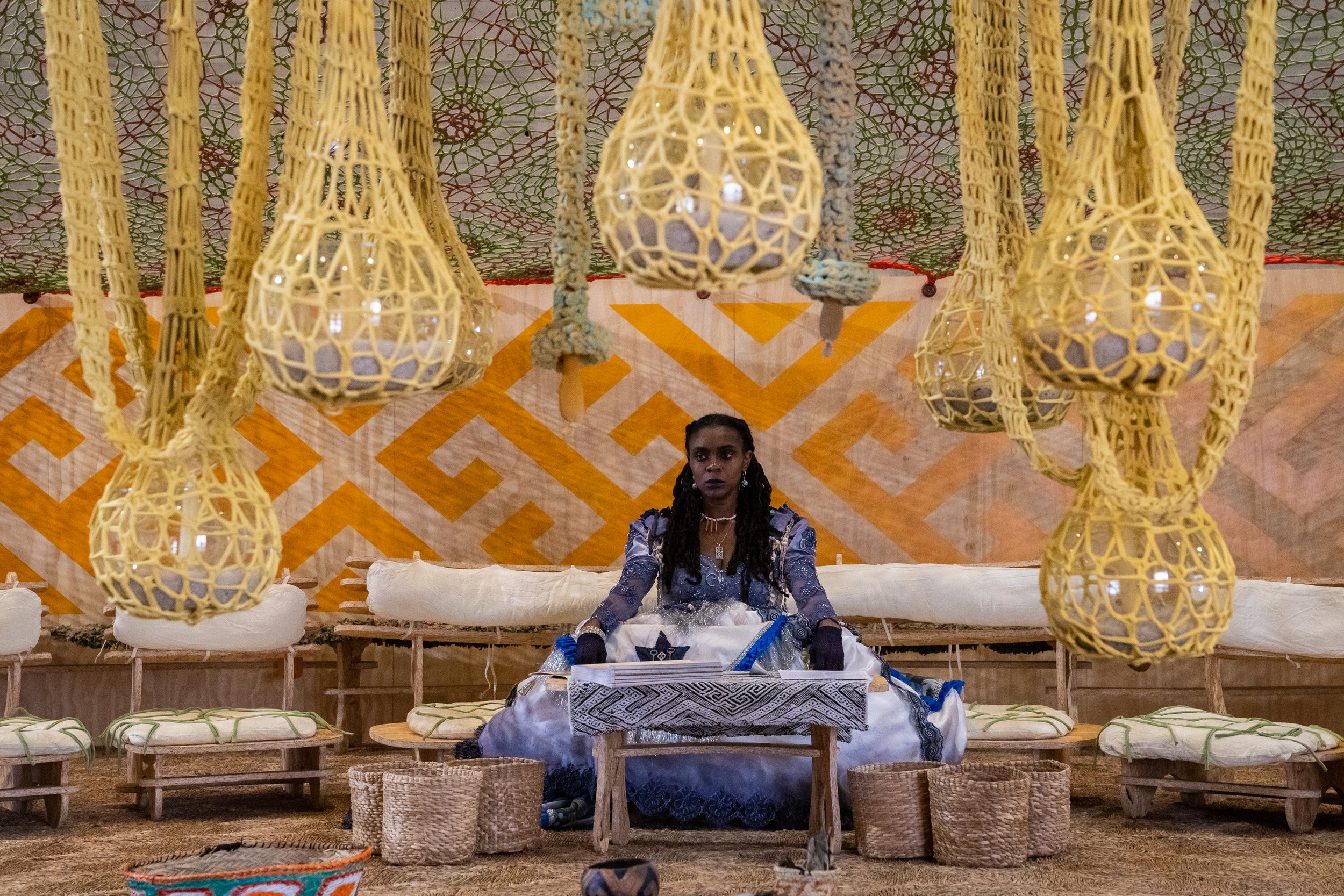 A woman sits on a bench wearing blue and white regalia. She is surrounded my woven baskets and lamps and large yellow-and-white quilted textiles.