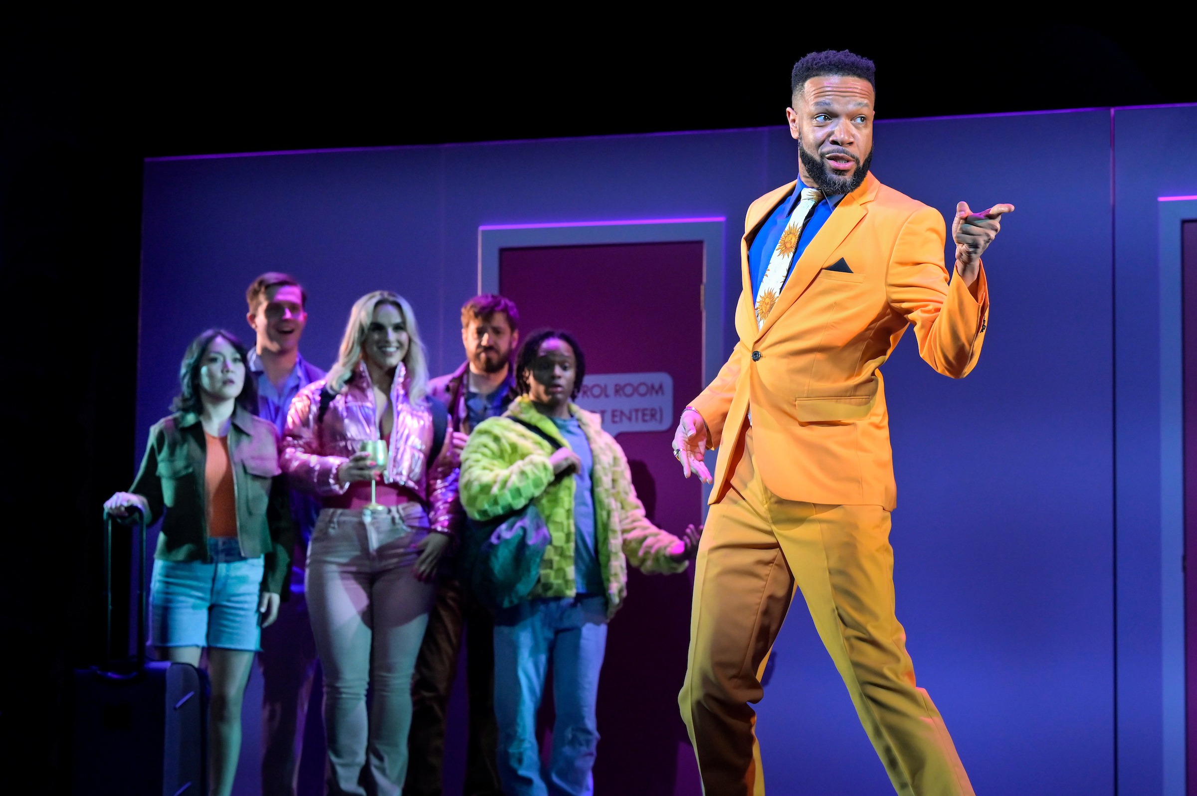 A performer in a bright orange suit stands on a stage under colorful lighting, striking a confident pose and pointing outward. Behind him, a small group of casually dressed people watch with mixed expressions—some smiling, some surprised—near a doorway labeled “Control Room.” The scene has a theatrical, lively atmosphere with bold colors and dramatic lighting.