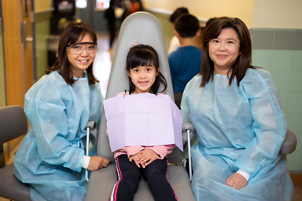 A child patient poses in a dental chair with a dentist and hygienist