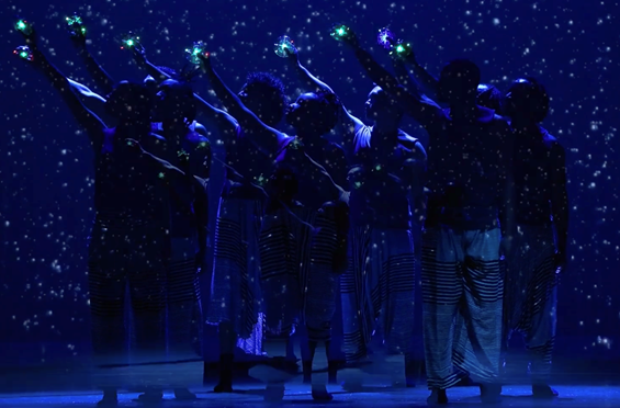 A group of seven dancers are standing on a dark stage. The outline of their bodies is faintly lit with blue light. Their right arms are lifted up and it looks like they are holding stars.