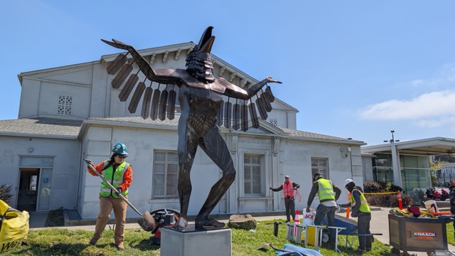 Workers install a large winged human-bird sculpture outside a building, using tools and safety gear under clear skies.