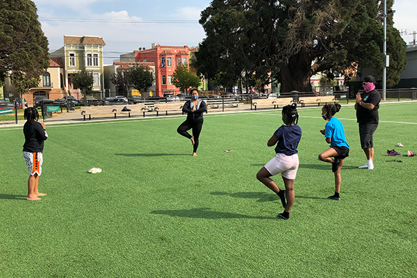 Young people practice balancing on one foot in an AstroTurf green in a park.