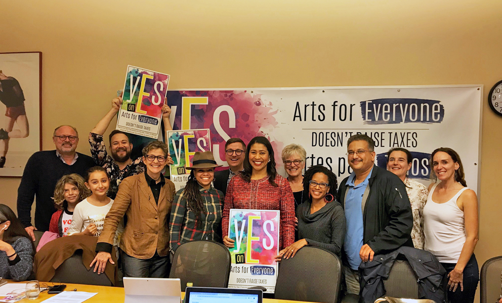 Group of people smiling indoors, holding “YES on Arts for Everyone” signs in front of a campaign banner.